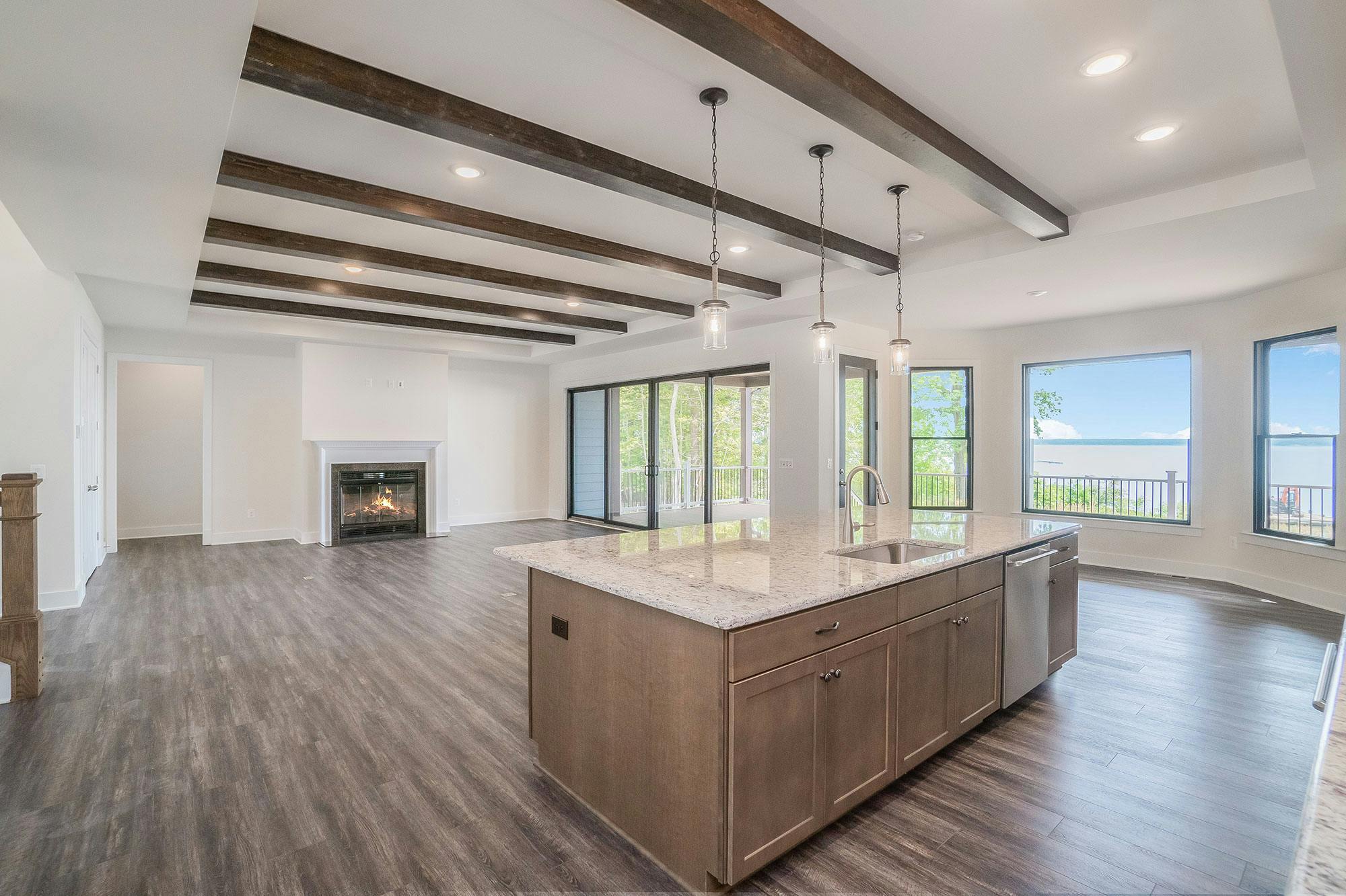 Large open room with a high ceiling and wood accent beams.  A fireplace is at one end of the room and a wood kitchen island with a white marble counter overlooks a view of the water with bay windows and sliding door.